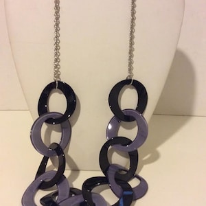 Puede incluir: Un collar de cadena plateada con eslabones grandes de plástico negro y morado. Los eslabones son de forma ovalada y tienen un acabado brillante.