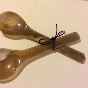 May include: A set of two horn salad servers tied together with a black ribbon. The servers are made of natural horn and have a light brown color.
