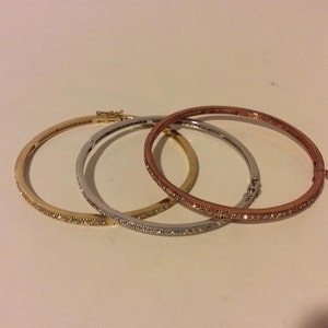 Può includere: Tre braccialetti rigidi con un design scintillante di cristalli trasparenti. I braccialetti sono in oro, argento e oro rosa.