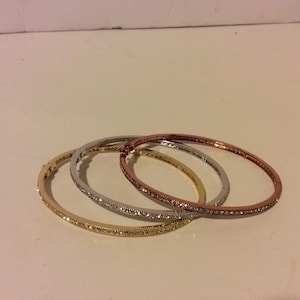 Può includere: Tre braccialetti in oro, argento e oro rosa, ognuno con una fila di strass trasparenti attorno al bordo.