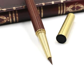 May include: A wooden calligraphy pen with a gold tip and a matching gold cap. The pen is lying on a white surface next to a closed book with a brown leather cover.