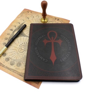 Op de afbeelding: Een zwart lederen gebonden dagboek met een rood ankh-symbool en de tekst "Campaign Journal Historical Events". Het dagboek ligt op een vintage kaart met een houten stempel en een gouden basis.