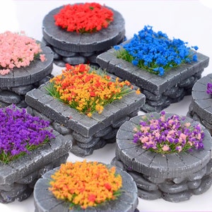 May include: A selection of miniature grey stone structures, each with a different colour of artificial flowers. The flowers are in shades of red, pink, blue, orange, purple, and a mix of pink and purple. The structures are square and round.
