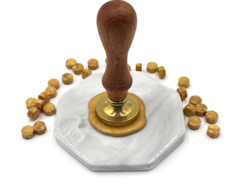 May include: A wooden handle wax seal stamp with a brass base and a gold wax seal on a white marble base. The stamp is surrounded by gold wax seal pellets.