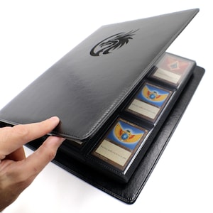 May include: Black card binder with a dragon logo, holding trading cards. The binder is open, revealing several cards in protective sleeves. The cards have colorful illustrations and text. The binder is made of a smooth, leather-like material.