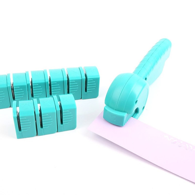 May include: A set of turquoise blue plastic corner rounder tools for paper crafts. The tools are designed to round the corners of paper, cards, and other materials. The tools are shown with a pink piece of paper.