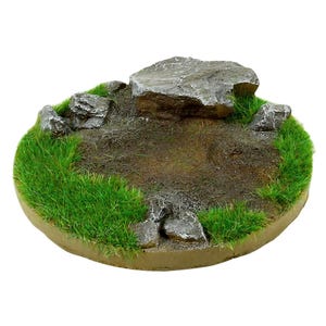 Op de afbeelding: Een miniatuur terreinbasis met een grote grijze rotsformatie, omringd door felgroen gras en kleinere grijze rotsen. De basis heeft een cirkelvorm met een bruin, aardeachtig centrum en een beige rand, ideaal voor tabletop games.