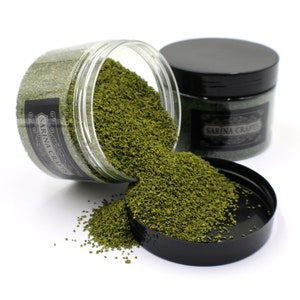 May include: Two jars of green craft sand, one jar is tipped over and spilling the sand. The jars have black lids and labels that say "Sarina Crafts".