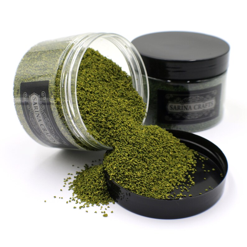 May include: Two jars of green craft sand, one jar is tipped over and spilling the sand. The jars have black lids and labels that say "Sarina Crafts".