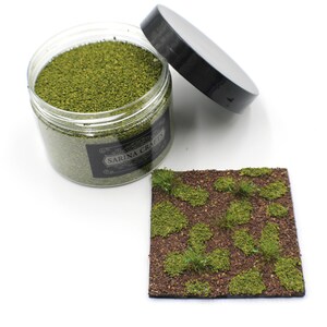 May include: A clear jar filled with green model train landscaping material. The jar has a black label with the text "SARINA CRAFTS". A small square of brown material with green tufts of grass is shown next to the jar.