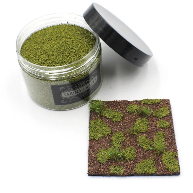 May include: A clear jar filled with green model train landscaping material. The jar has a black label with the text "SARINA CRAFTS". A small square of brown material with green tufts of grass is shown next to the jar.