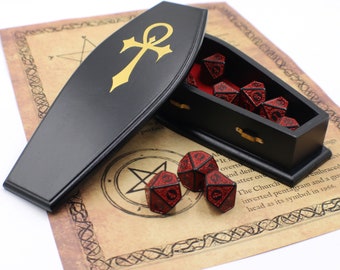 May include: A black wooden coffin-shaped box with a gold ankh symbol on the lid. The box is open and contains red and black dice with a metallic finish. The box is sitting on a parchment paper with a pentagram and other symbols.
