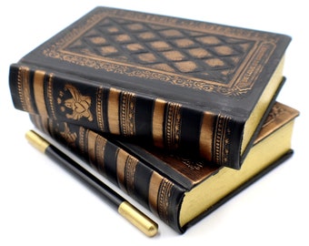 May include: Two black leather-bound journals with gold accents and a gold pen. The journals have a decorative pattern on the cover and are stacked on top of each other.