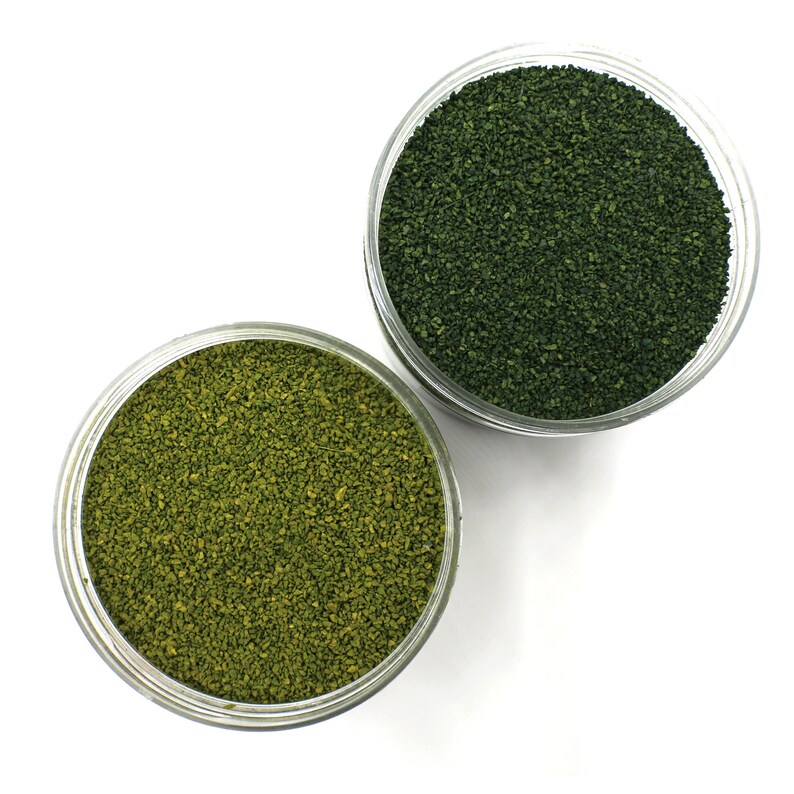 May include: Two clear glass jars filled with green and light green granular material. The jars are on a white background.
