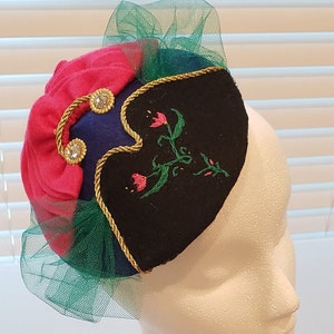 May include: A black felt hat with a green and pink tulle trim. The hat has a gold braid trim and embroidered flowers. The hat is on a white mannequin head. A picture frame with a picture of two girls in dresses is in the background.