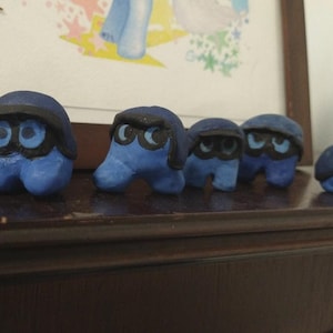 Puede incluir: Una colección de cinco figuras de arcilla azul con detalles negros. Cada figura tiene un cuerpo redondeado, ojos grandes y una cubierta oscura para la cabeza. Están dispuestas sobre una superficie de madera oscura, con una imagen enmarcada en el fondo.