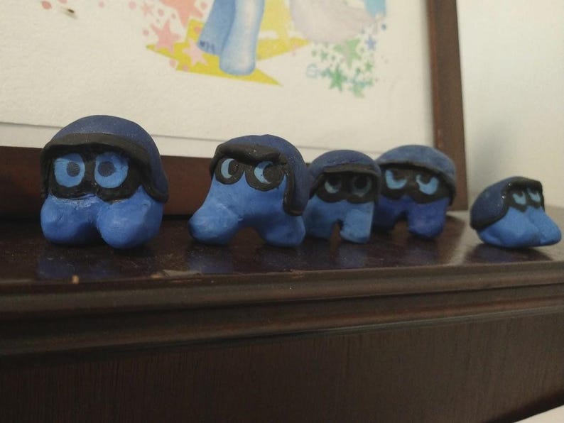 Puede incluir: Una colecci&oacute;n de cinco figuras de arcilla azul con detalles negros. Cada figura tiene un cuerpo redondeado, ojos grandes y una cubierta oscura para la cabeza. Est&aacute;n dispuestas sobre una superficie de madera oscura, con una imagen enmarcada en el fondo.