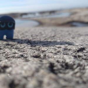 Puede incluir: Una peque&ntilde;a figura azul con una m&aacute;scara negra y ojos turquesa se encuentra sobre una superficie gris texturizada. La figura tiene una cabeza redondeada y un cuerpo rectangular con una peque&ntilde;a abertura en la parte inferior. El fondo est&aacute; borroso, con un indicio de agua y cielo.
