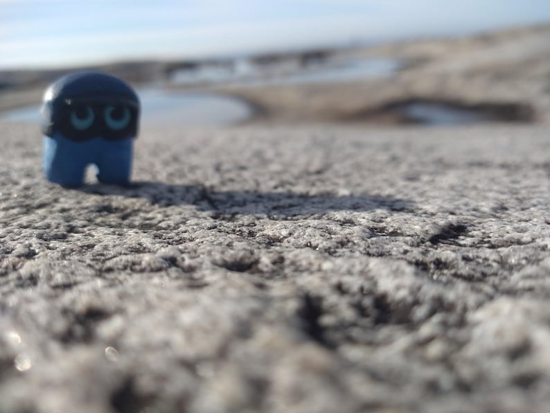 Puede incluir: Una peque&ntilde;a figura azul con una m&aacute;scara negra y ojos turquesa se encuentra sobre una superficie gris texturizada. La figura tiene una cabeza redondeada y un cuerpo rectangular con una peque&ntilde;a abertura en la parte inferior. El fondo est&aacute; borroso, con un indicio de agua y cielo.