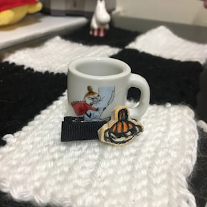May include: A small white ceramic mug with a black and white illustration of a character from the Moomin series. The mug is sitting on a white and black crocheted surface. There is a small wooden ornament with a pumpkin design next to the mug.