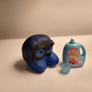 Puede incluir: Una escultura azul estilizada de un personaje con casco y gafas negros, junto a una botella de detergente para ropa azul claro con una etiqueta rosa y las palabras "Laundry Detergent". Tambi&eacute;n hay un peque&ntilde;o objeto azul claro en forma de estrella.