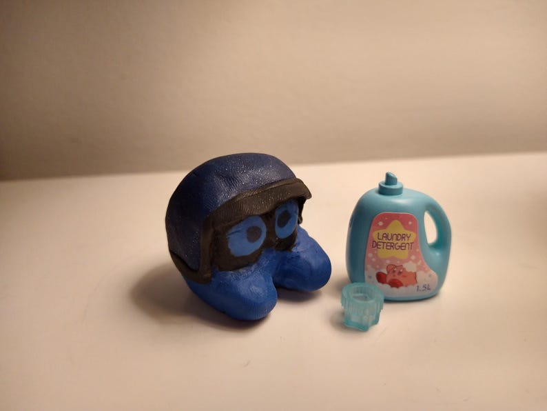 Puede incluir: Una escultura azul estilizada de un personaje con casco y gafas negros, junto a una botella de detergente para ropa azul claro con una etiqueta rosa y las palabras "Laundry Detergent". Tambi&eacute;n hay un peque&ntilde;o objeto azul claro en forma de estrella.