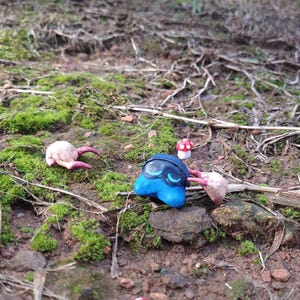 Puede incluir: Una escena caprichosa con figuras de arcilla hechas a mano. Una criatura azul con detalles negros y otras dos con garras rosas est&aacute;n dispersas entre musgo y ramitas. Una peque&ntilde;a seta roja y blanca a&ntilde;ade un toque de fantas&iacute;a al entorno natural.