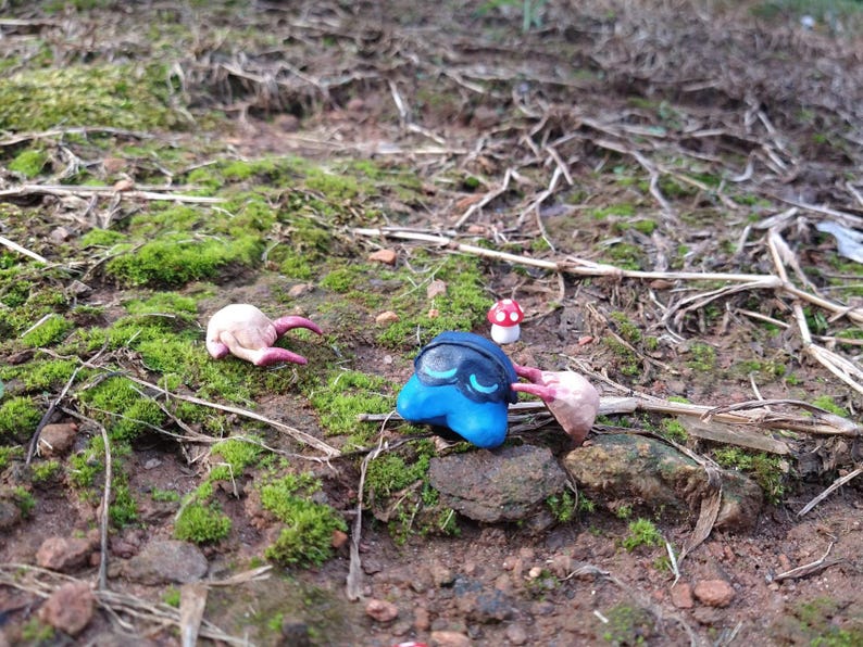 Puede incluir: Una escena caprichosa con figuras de arcilla hechas a mano. Una criatura azul con detalles negros y otras dos con garras rosas est&aacute;n dispersas entre musgo y ramitas. Una peque&ntilde;a seta roja y blanca a&ntilde;ade un toque de fantas&iacute;a al entorno natural.
