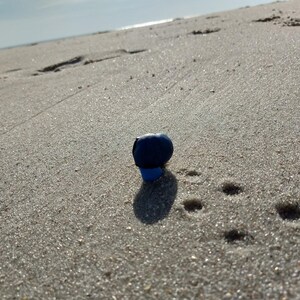 Puede incluir: Un objeto azul oscuro, posiblemente un tap&oacute;n de botella, descansa sobre una playa arenosa. La sombra del objeto es visible en la arena, con huellas en el fondo. La escena est&aacute; iluminada por la luz del sol.