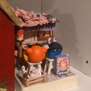 Puede incluir: Escena en miniatura de una tienda japonesa con techo de flores de cerezo y figuras de dibujos animados. Un letrero dice "Happiness". La escena incluye una caja de madera y peque&ntilde;os taburetes, creando un diorama caprichoso.