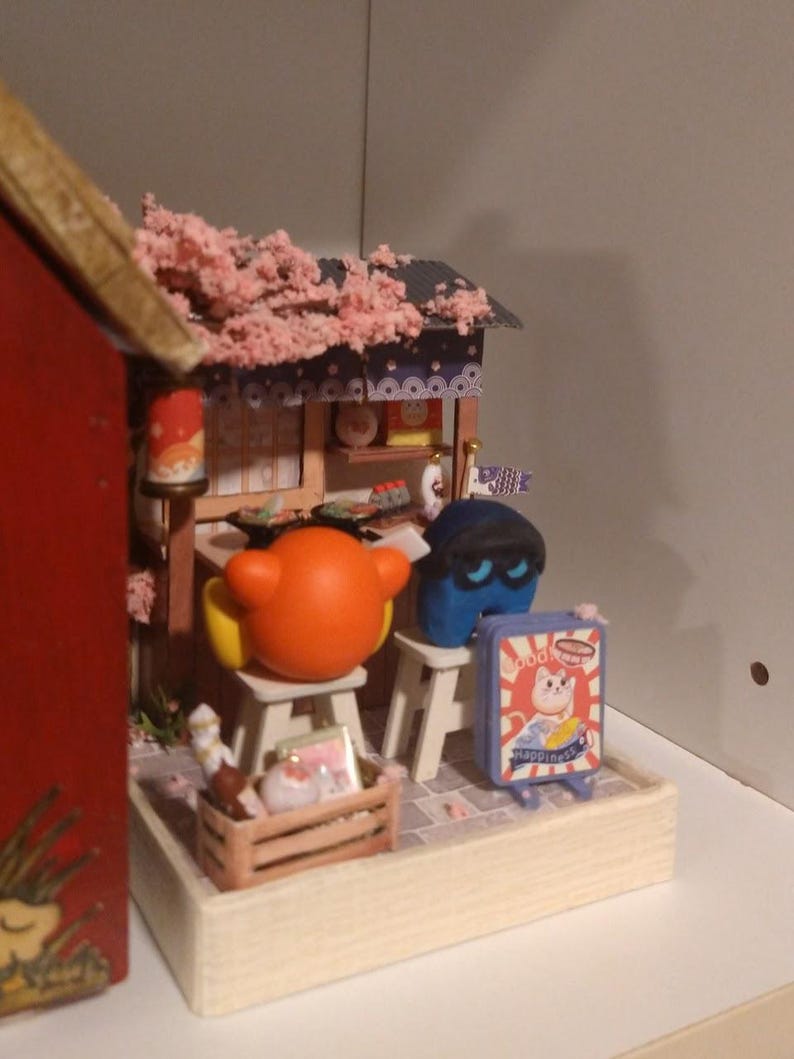 Puede incluir: Escena en miniatura de una tienda japonesa con techo de flores de cerezo y figuras de dibujos animados. Un letrero dice "Happiness". La escena incluye una caja de madera y peque&ntilde;os taburetes, creando un diorama caprichoso.