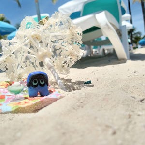 Puede incluir: Una escena playera caprichosa con una figura azul y negra de ojos grandes, una sombrilla de encaje blanco en miniatura y una alfombra con estampado floral. La escena est&aacute; ambientada en una playa de arena con tumbonas y sombrillas al fondo.