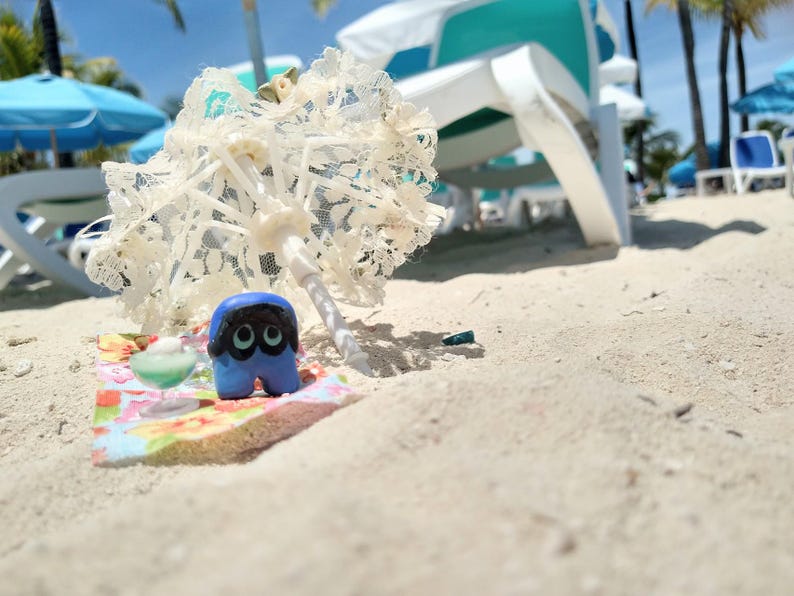 Puede incluir: Una escena playera caprichosa con una figura azul y negra de ojos grandes, una sombrilla de encaje blanco en miniatura y una alfombra con estampado floral. La escena est&aacute; ambientada en una playa de arena con tumbonas y sombrillas al fondo.