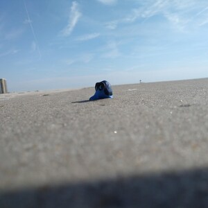 Puede incluir: Una peque&ntilde;a escultura azul con detalles negros descansa sobre una playa arenosa bajo un cielo azul brillante. La escultura es el foco principal, con el mar y los edificios al fondo. La imagen est&aacute; tomada desde un &aacute;ngulo bajo.