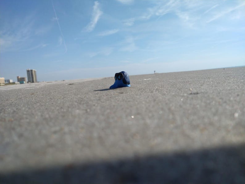 Puede incluir: Una peque&ntilde;a escultura azul con detalles negros descansa sobre una playa arenosa bajo un cielo azul brillante. La escultura es el foco principal, con el mar y los edificios al fondo. La imagen est&aacute; tomada desde un &aacute;ngulo bajo.