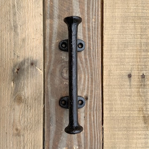 May include: Black cast iron door handle with a rounded top and two oval-shaped plates. The handle is mounted on a wooden surface.