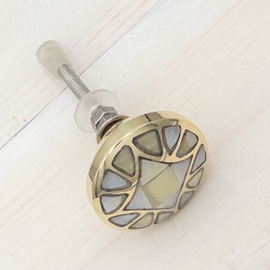 May include: A round, brass cabinet knob with a geometric design. The knob is made of brass and has a white, mother-of-pearl inlay. The knob is attached to a screw.