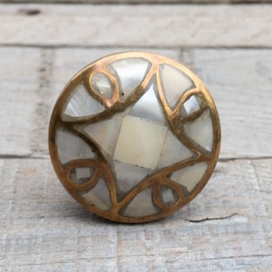 May include: Circular brass knob with a mother-of-pearl inlay. The knob has a geometric pattern with a central square and surrounding shapes. The brass has a warm, aged look, and the inlay has a pearlescent sheen.