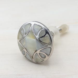 May include: A decorative bottle stopper with a silver-colored metal base and a round, inlaid design. The inlaid design features a star-like pattern with iridescent mother-of-pearl segments. The stopper has a clear, plastic-like stem.