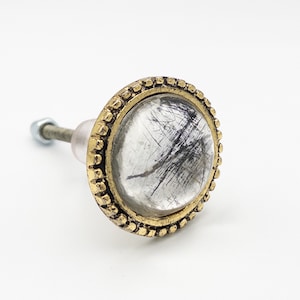 Peut inclure: Un bouton de meuble décoratif avec un centre en verre clair et rond contenant des inclusions noires. Le verre est serti dans un cadre en métal doré avec un bord perlé. Le bouton a une vis argentée.