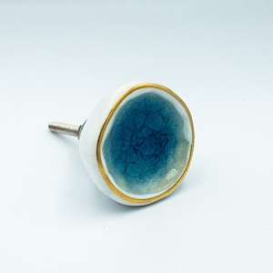 May include: A round cabinet knob with a white exterior and a concave center featuring a blue crackle glaze finish. The edge of the knob is trimmed with a thin gold-colored metallic band, and a threaded metal bolt extends from the back side.