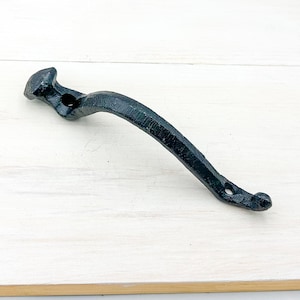 May include: A black, curved metal handle with a textured surface. The handle has a nail-like head on one end and a rounded end with a hole on the other. It is likely used for a cabinet or drawer.