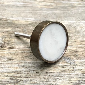 以下が含まれることがあります： 白い大理石の中心とブロンズ色の金属リムを備えた丸いキャビネットノブ。ノブには、取り付け用のねじ付きシルバーメタルステムが付いています。シンプルでエレガントなデザインで、さまざまな家具スタイルに適しています。