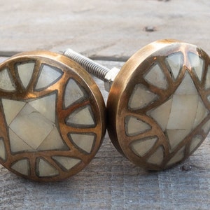 May include: Two round brass cabinet knobs with a white bone inlay design. The knobs have a geometric pattern with a square in the centre.