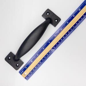 May include: A black cast iron door pull with a rounded handle and rectangular mounting plates. A blue and yellow ruler is next to the door pull. The ruler measures in inches.
