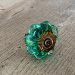 Peut inclure: Un bouton de meuble en verre vert clair facetté avec une base en métal de couleur bronze. Le bouton a un motif en forme d'étoile et est posé sur une surface en bois. Cette quincaillerie décorative ajoute une touche d'élégance aux meubles.