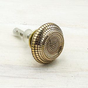 May include: A decorative brass cabinet knob with a textured, dotted surface. The knob has a circular design with a central pattern of smaller dots, surrounded by larger dots. The knob is attached to a clear, threaded post.