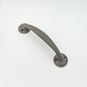 May include: A dark gray, curved door pull with two circular mounting plates. The handle has a slightly textured surface and a rustic, antique appearance. The door pull is made of metal and is designed for easy gripping.