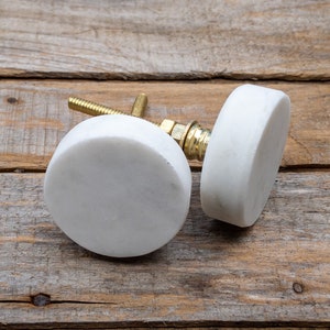 May include: Two white marble cabinet knobs with brass hardware. The knobs are round and have a smooth, polished surface. The brass hardware includes a threaded bolt and a nut for attaching to a cabinet door or drawer.