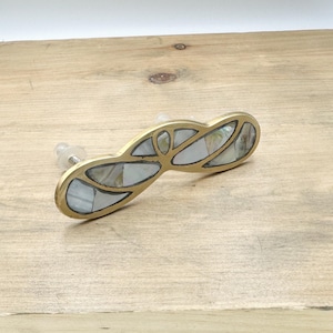 May include: A gold-toned cabinet pull with a unique design featuring inlaid mother-of-pearl pieces. The pull has a curved shape and is designed to be used on cabinets or drawers.